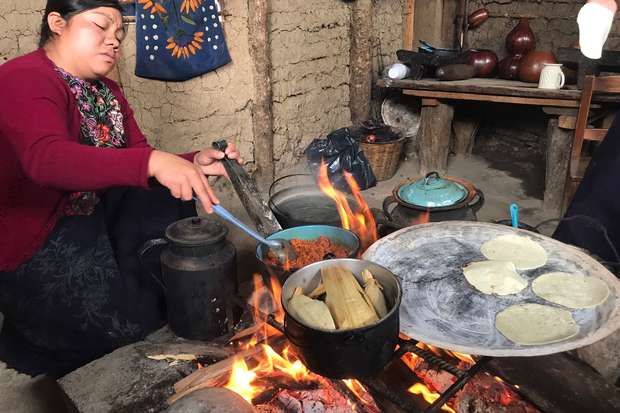 A person cooks over an open fire, preparing tortillas and tamales in a rustic kitchen with earthen walls and wooden shelves holding pottery items.